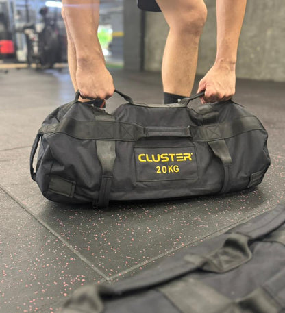 Person holding a black sand bag labeled 'Cluster 20 KG' in a gym setting.